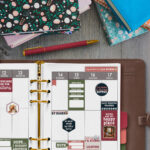 Flat lay of an open planner decorated with book-themed stickers surrounded by colorful books, a patterned notebook, and a red pen on a wooden table.