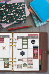 Flat lay of an open planner decorated with book-themed stickers surrounded by colorful books, a patterned notebook, and a red pen on a wooden table.