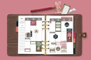 Open planner decorated with cozy book-themed stickers&mdash;including reading quotes, labels, and trackers&mdash;set inside a brown planner cover on a pink background, with additional book-themed stickers and a red pen placed above the planner.