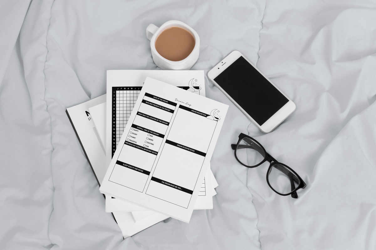 Dream tracker and dream log pages resting on rumpled bedding alongside a cup of coffee, glasses, and a smartphone, creating a cozy morning journaling scene.