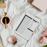 Flatlay of the dream tracker page on a white bedspread, surrounded by a pink mug with coffee, a heart-shaped dish, pink roses, a journal, and a perfume bottle.