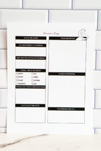 Printed dream log page displayed against a white subway-tile wall, showing sections for dream details, vibe checkboxes, notes, dream interpretation, and mood reflections.