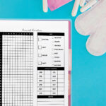 Flat lay of a yearly period tracker planner page in a binder on a bright blue background. The layout includes a daily grid, symptom key, cycle length chart, and notes section. Surrounding the planner are pink pad wrappers, tampons, a calendar with circled dates, and sanitary pads.