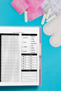 Flat lay of a yearly period tracker planner page in a binder on a bright blue background. The layout includes a daily grid, symptom key, cycle length chart, and notes section. Surrounding the planner are pink pad wrappers, tampons, a calendar with circled dates, and sanitary pads.
