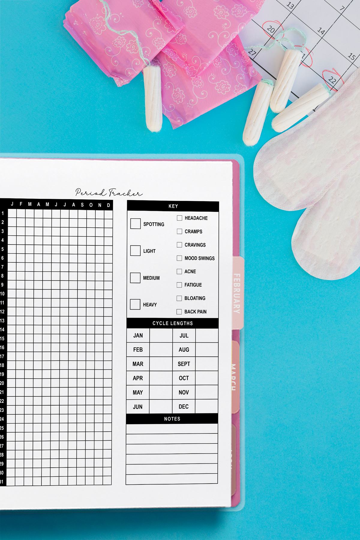 Flat lay of a yearly period tracker planner page in a binder on a bright blue background. The layout includes a daily grid, symptom key, cycle length chart, and notes section. Surrounding the planner are pink pad wrappers, tampons, a calendar with circled dates, and sanitary pads.