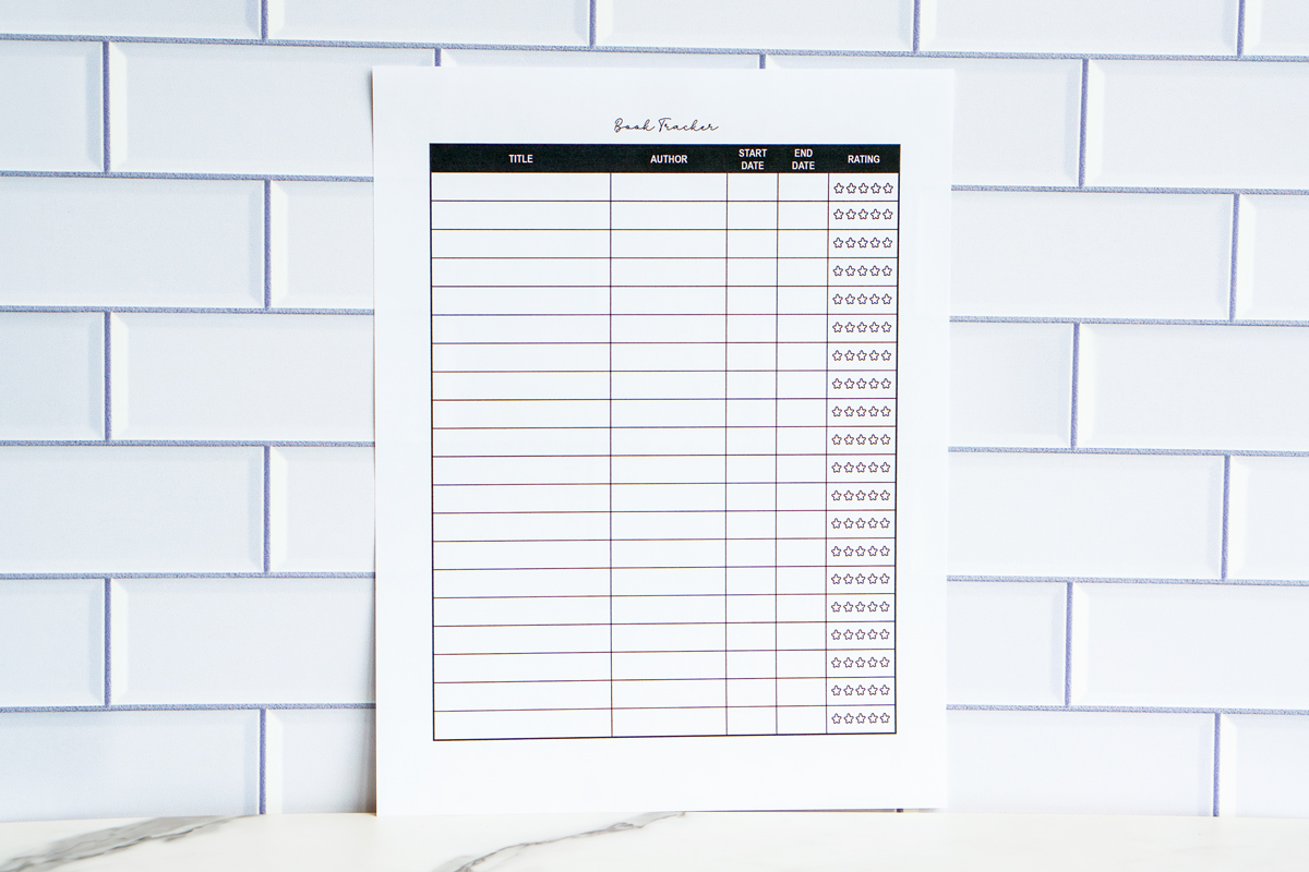 Horizontal image of a clean, black-and-white book tracker printable with columns for title, author, dates, and star ratings, photographed against a white subway-tile background.