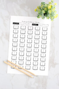 Printed 50 Books Reading Challenge tracker page displayed on a marble surface with two wooden pencils and a small potted plant, featuring rows of open book icons and spaces for start and end dates.
