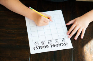A child writing on a &ldquo;School Goals Bingo&rdquo; sheet while seated at a dark wooden table, filling in the blank bingo squares with a pencil.