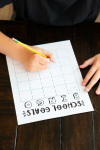 A child writing on a &ldquo;School Goals Bingo&rdquo; sheet with a pencil at a dark wooden table, filling in blank squares on the bingo grid.