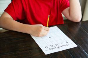 A child in a red shirt sitting at a wooden table writing on a &ldquo;Personal Goals Bingo&rdquo; sheet, holding a yellow pencil over the blank bingo grid with a star in the center.