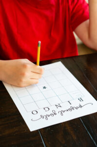 A close-up of a child in a red shirt writing on a &ldquo;Personal Goals Bingo&rdquo; page at a dark wooden table, with a pencil poised above the center star square.