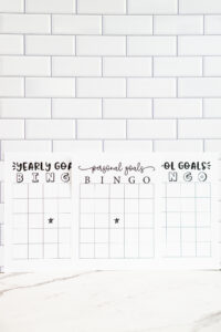 A wide vertical photo of three goal-themed bingo sheets&mdash;Yearly, Personal, and School Goals Bingo&mdash;lined up along a white subway tile wall with a marble surface below.