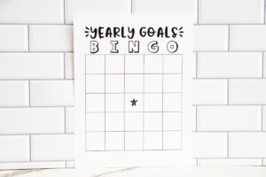 A &ldquo;Yearly Goals Bingo&rdquo; printable sheet displayed upright against a white subway tile wall, showing a blank bingo grid with a star in the middle.