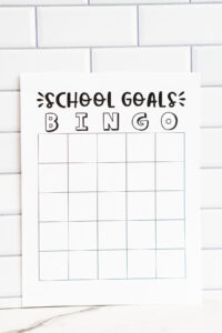 A &ldquo;School Goals Bingo&rdquo; printable sheet photographed upright against a white subway tile background, showing an empty bingo grid with no star in the center.