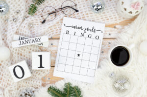 A styled flat lay featuring a &ldquo;Career Goals Bingo&rdquo; printable on a rustic wooden surface, surrounded by cozy winter d&eacute;cor including a white knit blanket, calendar blocks showing January 01, a cup of coffee, ornaments, and evergreen sprigs.