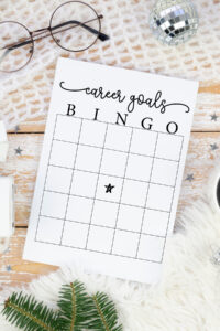 A close-up overhead view of a &ldquo;Career Goals Bingo&rdquo; sheet on a wooden surface with winter-themed props such as eyeglasses, a disco ball ornament, star confetti, and a cup of coffee.