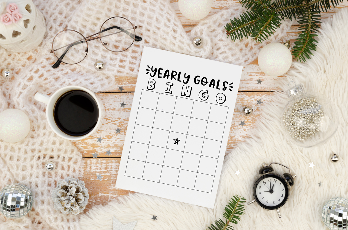 A styled flat lay of a &ldquo;Yearly Goals Bingo&rdquo; printable on a weathered wooden background, surrounded by winter decorations including a fuzzy white blanket, coffee cup, ornaments, pinecones, and an alarm clock.