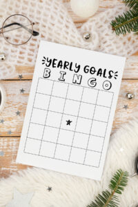 A close-up overhead photo of a &ldquo;Yearly Goals Bingo&rdquo; sheet placed on a wooden surface with winter d&eacute;cor, including a knit blanket, ornaments, star confetti, and evergreen branches.