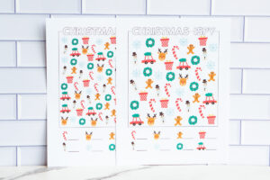Two Christmas I-Spy worksheets displayed upright against a white tile wall. Each sheet includes colorful holiday icons like wreaths, reindeer, gingerbread men, and snowflakes.