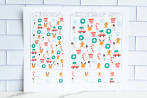 Two printed Christmas I-Spy worksheets displayed side by side, each filled with colorful holiday icons like candy canes, wreaths, reindeer faces, snowmen, snowflakes, presents, and cars with Christmas trees.
