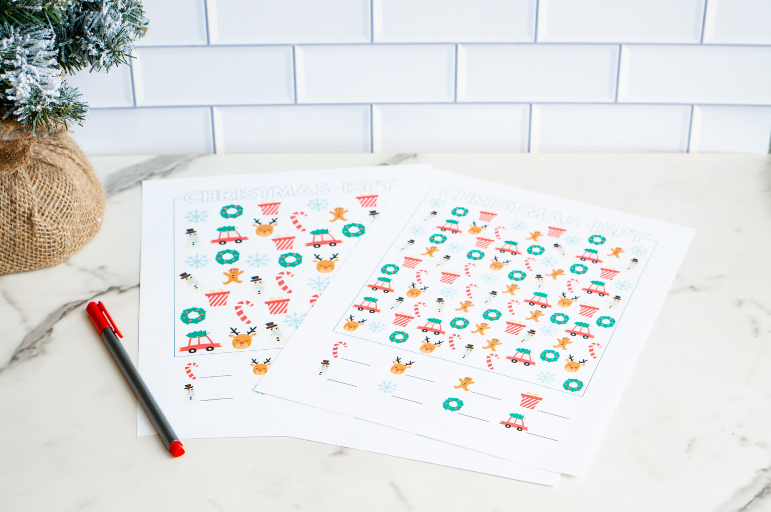 Two Christmas I-Spy printable pages on a marble surface with a red-tipped pen beside them. The pages feature colorful holiday icons like wreaths, reindeer, candy canes, snowmen, and presents.