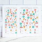 Two printed Christmas I-Spy worksheets displayed side by side, each filled with colorful holiday icons like candy canes, wreaths, reindeer faces, snowmen, snowflakes, presents, and cars with Christmas trees.