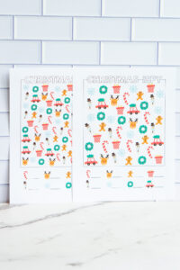 Two printed Christmas I-Spy worksheets displayed side by side, each filled with colorful holiday icons like candy canes, wreaths, reindeer faces, snowmen, snowflakes, presents, and cars with Christmas trees.