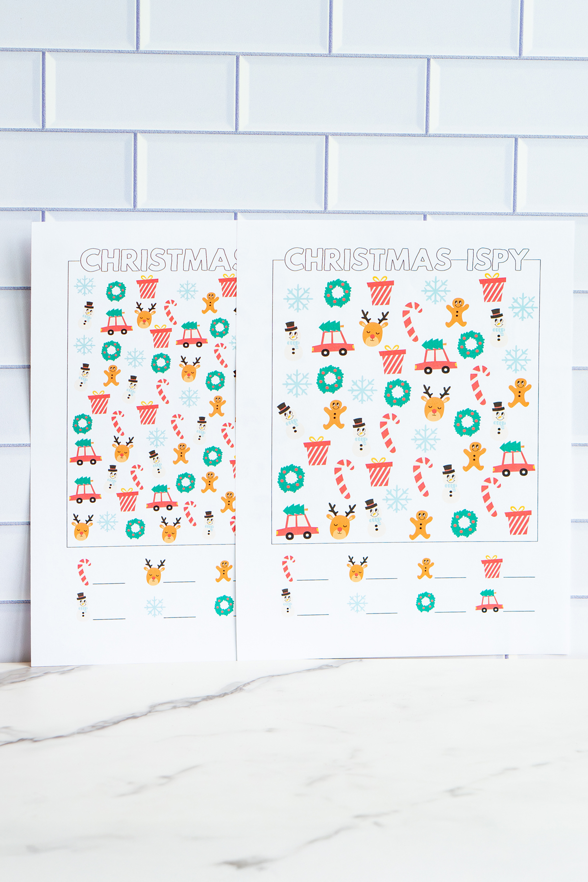 Two printed Christmas I-Spy worksheets displayed side by side, each filled with colorful holiday icons like candy canes, wreaths, reindeer faces, snowmen, snowflakes, presents, and cars with Christmas trees.