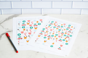 Two Christmas I-Spy printables lying on a marble surface beside a red pen, featuring colorful holiday icon patterns ready for kids to count.