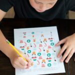 Child working on a printed Christmas I-Spy worksheet at a wooden table, counting and marking holiday icons with a pencil.