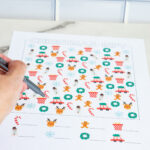 Close-up of a hand holding a pen while pointing at a colorful Christmas I-Spy worksheet laid on a marble surface.