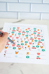 Close-up of a hand holding a pen while pointing at a colorful Christmas I-Spy worksheet laid on a marble surface.
