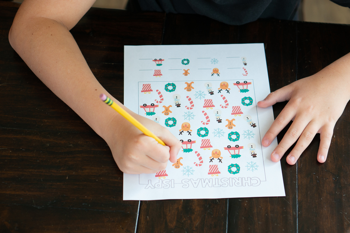 Child using a pencil to complete a Christmas I-Spy printable full of colorful holiday icons on a wooden table.