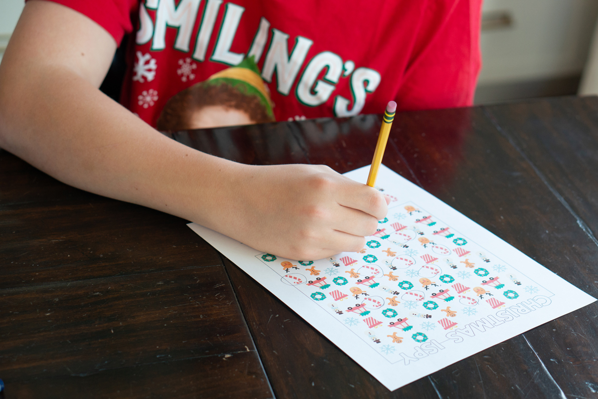 Child wearing a red Christmas shirt filling out a colorful Christmas I-Spy worksheet at a wooden table.