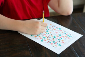 Child wearing a red Christmas shirt filling out a colorful Christmas I-Spy worksheet at a wooden table.