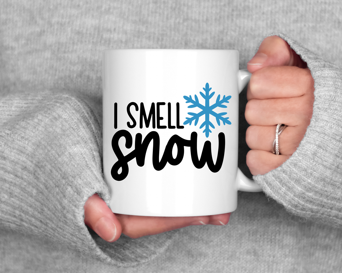 White mug held between two hands in cozy gray sweater sleeves, featuring the phrase ‘I Smell Snow’ with a blue snowflake graphic.