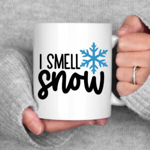 White mug held between two hands in cozy gray sweater sleeves, featuring the phrase &lsquo;I Smell Snow&rsquo; with a blue snowflake graphic.