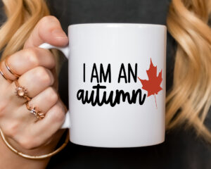 Close-up of a white ceramic mug held in a hand, featuring the phrase &lsquo;I Am an Autumn&rsquo; with a red maple leaf design.