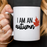 Close-up of a white ceramic mug held in a hand, featuring the phrase ‘I Am an Autumn’ with a red maple leaf design.