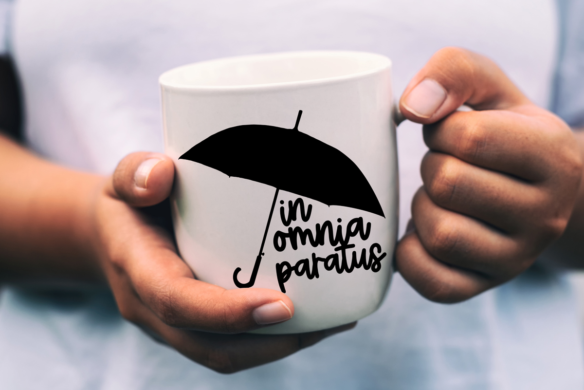 Close-up of hands holding a white mug printed with a black umbrella and the phrase ‘In Omnia Paratus.’