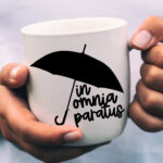 Close-up of hands holding a white mug printed with a black umbrella and the phrase ‘In Omnia Paratus.’