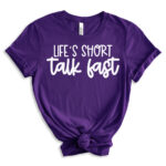 Purple short-sleeve t-shirt with the phrase “Life’s Short, Talk Fast” in bold white lettering, styled with rolled sleeves and a front knot.