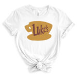 White t-shirt featuring a large golden coffee cup graphic with the word “Luke’s” in brown, inspired by the Gilmore Girls diner logo, styled with rolled sleeves and a front knot.