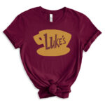 Burgundy t-shirt featuring a large golden coffee cup graphic with the word “Luke’s” in brown, inspired by the Gilmore Girls diner logo, styled with rolled sleeves and a front knot.