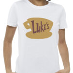 White T-shirt featuring the brown “Luke’s” diner coffee cup logo from Gilmore Girls, worn by a smiling woman.