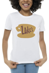 White T-shirt featuring the brown &ldquo;Luke&rsquo;s&rdquo; diner coffee cup logo from Gilmore Girls, worn by a smiling woman.