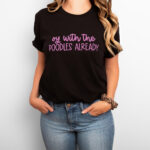 Woman wearing a black t-shirt that says “oy with the POODLES ALREADY” in purple lettering, paired with jeans and a leopard scrunchie.