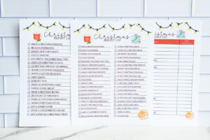 Three Christmas bucket list printables displayed side-by-side on a tile background: two full check-off lists and one customizable version with a date-completed column.
