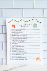 Christmas bucket list printable featuring illustrated string lights, hot cocoa, and ice skates, photographed upright against a tile background.