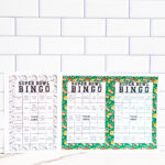 Three printed Super Bowl Bingo sheets&mdash;one black-and-white and two with green sports borders&mdash;standing upright against a white subway tile wall.